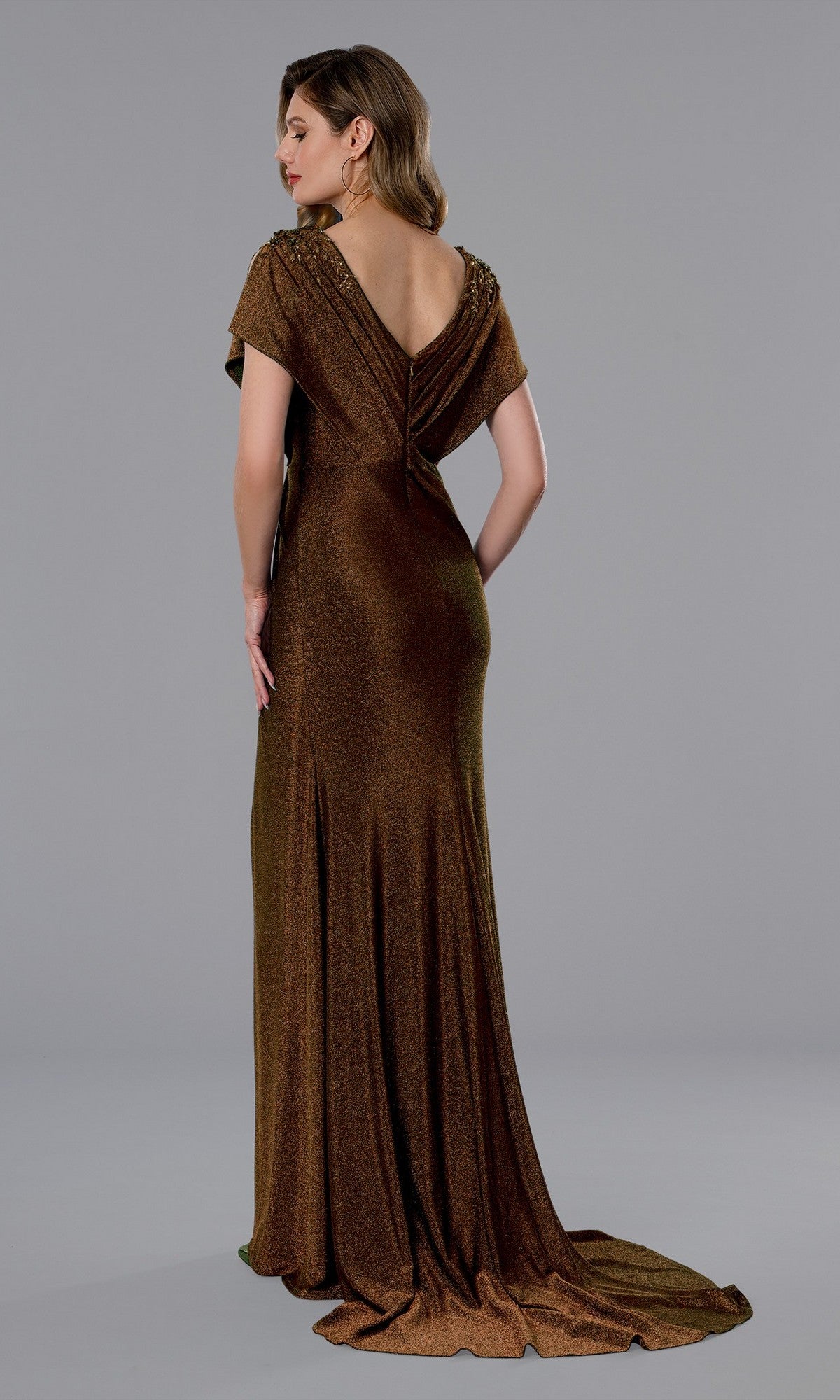 A woman poses against a plain gray background in the Stella Couture 25350 long formal dress, a floor-length metallic brown gown featuring short sleeves and a deep V-back.
